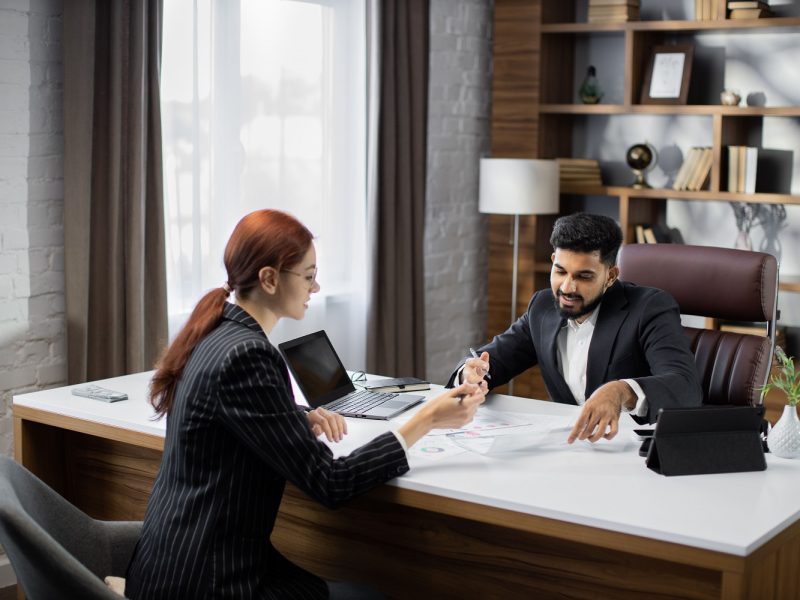 Handsome bearded businessman dealer showing papers with new agreement to his red hair businesswoman customer in a desktop, sitting at table in modern office.