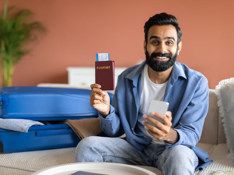 Tickets Booking Service. Cheerful Indian Traveler Man With Phone Showing His Passport With Boarding Pass At Home, Booking And Planning His Vacation Online Via New Travel Application