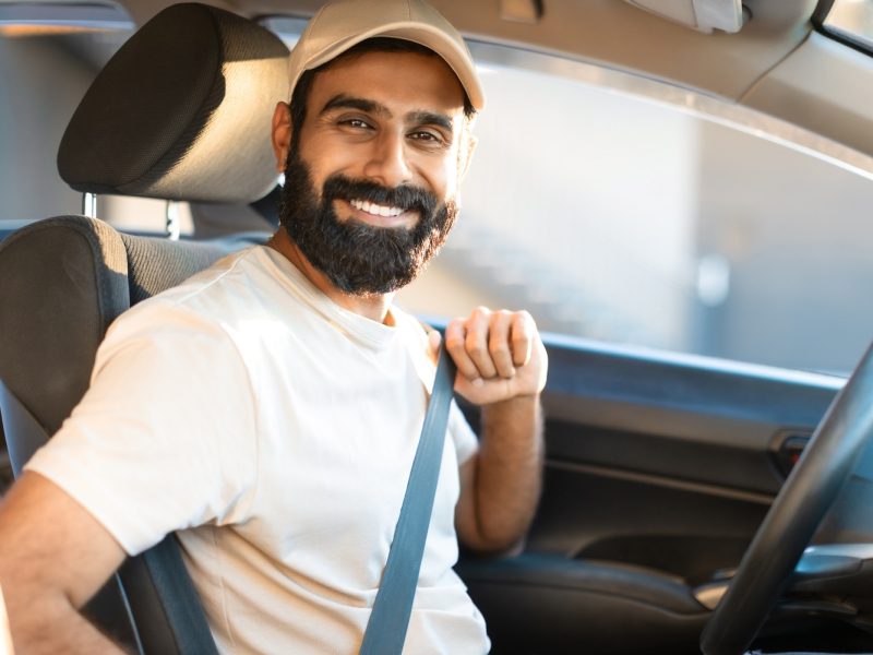 Cheerful Indian Young Man Sitting In Driver Seat In Car, Putting On Seat Belt For Safety On The Road During Travel By Auto, Posing Wearing Beige Cap And T-Shirt. Safe Vehicle, Insurance Offer
