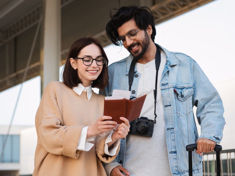 Cheerful young indian man and caucasian woman tourists in casual and glasses look at stamp in passport at airport, in city, outdoor. Start of travel, vacation and adventure together