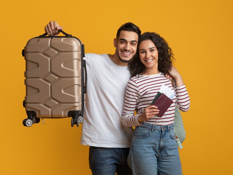 Family Trip. Happy Arabic Couple With Passports And Suitcase Posing Over Yellow Background, Joyful Middle Eastern Spouses Holding Luggage And Tickets, Ready For Travel Journey, Copy Space
