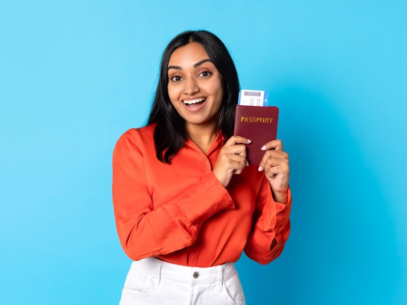 Happy young indian lady traveler showing passport and boarding pass in blue studio, smiling to camera and promoting travel offer for millennials, modern holiday advertisement banner