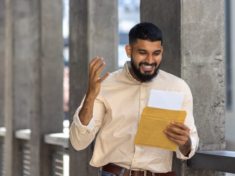 A cheerful bearded man stands outside, smiling widely as he reads a letter from a yellow envelope. The image captures a sense of joy, surprise, and positive emotion, ideal for concepts of good news, communication, and happiness.