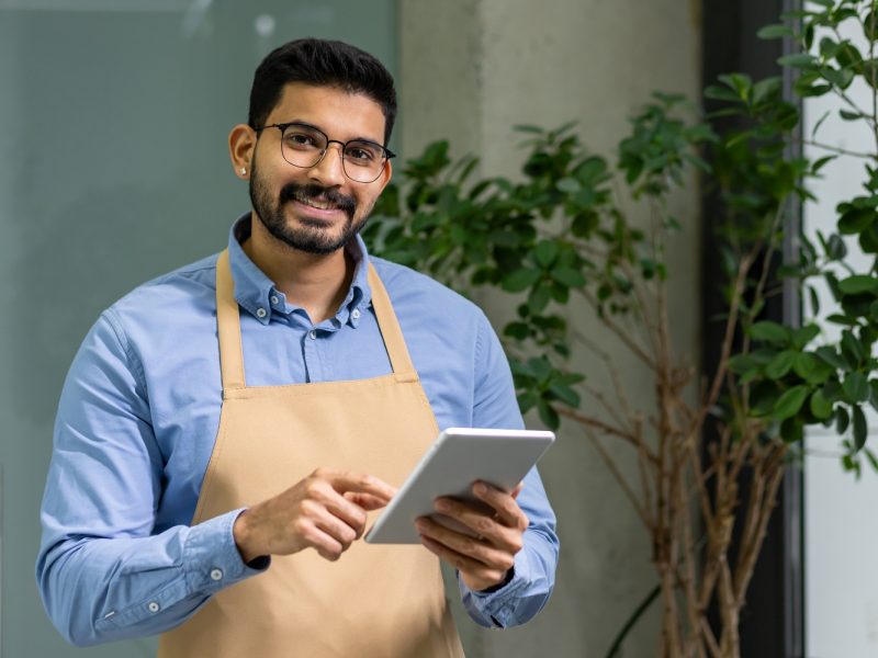 Cheerful male small business owner in apron operates tablet in green plant-filled contemporary workspace, exemplifying modern entrepreneurship.