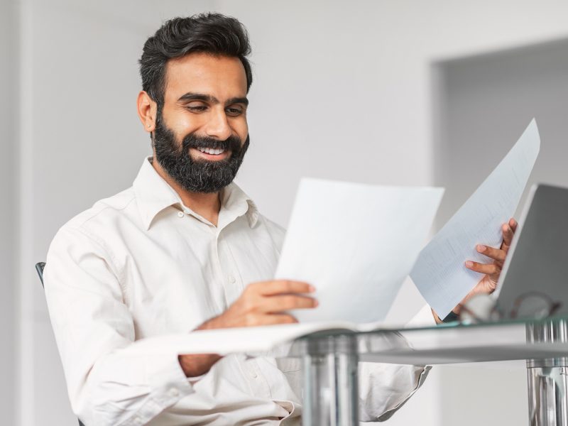 Cheerful indian businessman with beard happily examining documents, suggesting productive workflow or positive news at his workplace with laptop