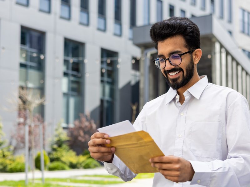 Cheerful bearded man in glasses and white shirt enjoying good news in a letter with office backdrop.