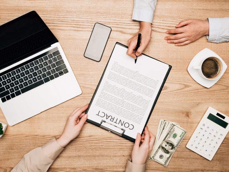Top view of business partners sign contract by laptop, dollar banknotes and coffee on table, cropped