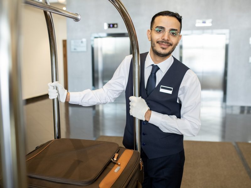 Young porter in uniform and eyeglasses pushing cart with suitcases of guests while moving in front of camera along corridor during work